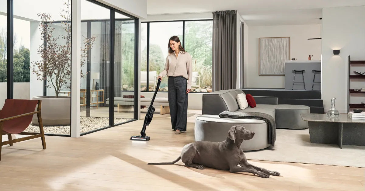 A woman vacuuming a bright living room while her dog rests on the floor